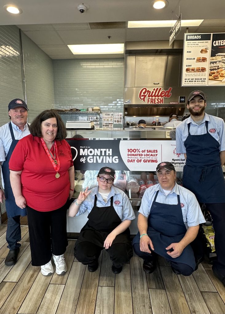 Jersey Mikes - Athlete and employees in front of "Month of Giving" sign